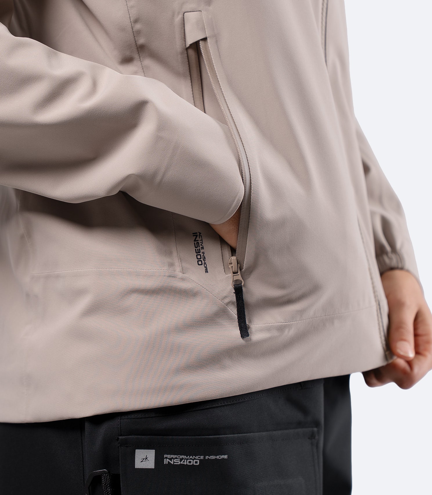 Primer plano de la mano en el bolsillo lateral de la chaqueta Zhik Youth INS300 Inshore Active Jacket en color piedra, mostrando la cremallera resistente al agua.